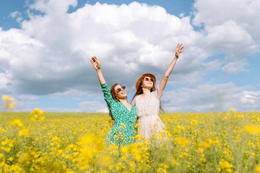 Şık giyinmiş iki neşeli kız arkadaş çiçek açan bir kolza tohumu tarlasında yürüyorlar. Yürüme, aktif yaşam tarzı, tatiller.