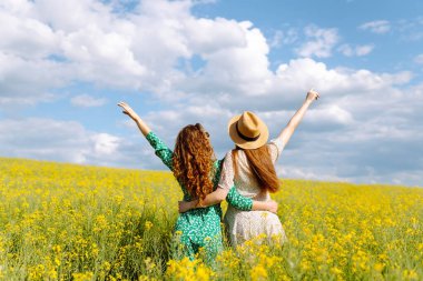 Şık giyinmiş iki neşeli kız arkadaş çiçek açan bir kolza tohumu tarlasında yürüyorlar. Yürüme, aktif yaşam tarzı, tatiller.