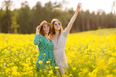 Şık giyinmiş iki neşeli kız arkadaş çiçek açan bir kolza tohumu tarlasında yürüyorlar. Yürüme, aktif yaşam tarzı, tatiller.