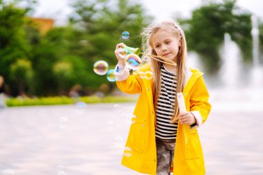 Sarı yağmurluklu neşeli küçük kız sabun köpükleri üflüyor, sonbaharda parkta yürüyüş yapıyor. Çocukluk, açık hava aktiviteleri, hafta sonları..