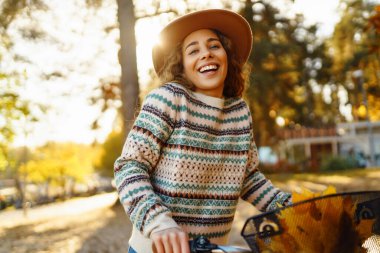 Şapkalı ve şık kazaklı gülümseyen genç bir kadın bisiklet yürüyüşü yapar ve günbatımında sarı yaprakların arasında sonbahar havasının tadını çıkarır..