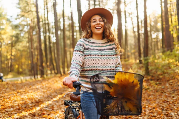帽子で笑顔の若い女性と自転車で歩くスタイリッシュなセーターは 日没に黄色い葉の中で森の中で秋の天気を楽しんでいます — ストック写真