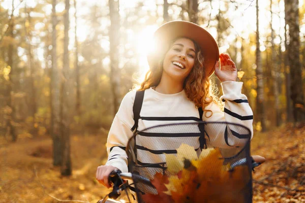 帽子で笑顔の若い女性と自転車で歩くスタイリッシュなセーターは 日没に黄色い葉の中で森の中で秋の天気を楽しんでいます — ストック写真