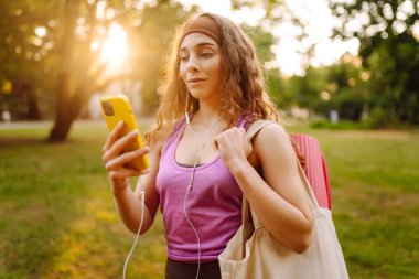 Spor çantalı güzel bir bayan güneşli bir parkta cep telefonuyla müzik dinliyor. Spor giyim bölümünde elinde telefon olan bir kadın. Spor konsepti, eğitim. Etkin yaşam biçimi.