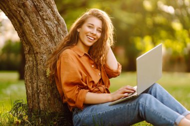 Dizüstü bilgisayarı olan sevimli genç bir kadın parktaki çimlerin üzerinde oturuyor. Çevrimiçi eğitim, uzaktan çalışma. Bağımsızlık, blog, iş konsepti.