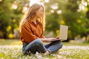 Dizüstü bilgisayarı olan sevimli genç bir kadın parktaki çimlerin üzerinde oturuyor. Çevrimiçi eğitim, uzaktan çalışma. Bağımsızlık, blog, iş konsepti.