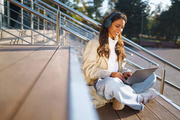 Happy woman in stylish clothes with laptop and headphones in the city works. Young woman listening to music, video conference or podcast. Lifestyle. Freelancing concept, online education.