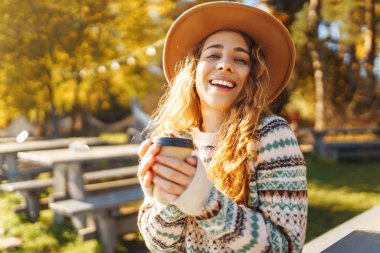 Şapkalı şirin bir kadının portresi göle bakan bir fincan kahveyle oturuyor. Kıvırcık bir kadının sonbahar parkında sıcak içkinin keyfini çıkarırken çekilmiş açık hava fotoğrafı..