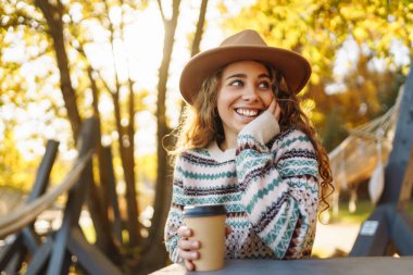Şapkalı şirin bir kadının portresi göle bakan bir fincan kahveyle oturuyor. Kıvırcık bir kadının sonbahar parkında sıcak içkinin keyfini çıkarırken çekilmiş açık hava fotoğrafı..