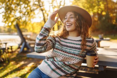 Şapkalı şirin bir kadının portresi göle bakan bir fincan kahveyle oturuyor. Kıvırcık bir kadının sonbahar parkında sıcak içkinin keyfini çıkarırken çekilmiş açık hava fotoğrafı..