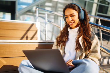 Dizüstü bilgisayarı ve kulaklığı olan normal giyinmiş, çalışan gülümseyen bir kız öğrenci. Dışarıda kablosuz kulaklıkları ve dizüstü bilgisayarı olan genç bir kadın..