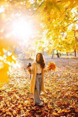 Sonbahar parkında, şık kıyafetler içinde mutlu bir kadın ve kamera düşmüş yaprakların arasında eğleniyor. Gülümseyen turist kadın, sonbahar mevsiminde sıcak ve güneşli havanın tadını çıkarıyor..