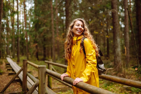 stock image Female tourist in a yellow coat with a backpack traveling along a wooden path in the forest. Trekking trail. Concept of travel, nature, vacation.