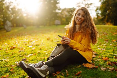 Elinde akıllı telefonuyla neşeli bir kadın sonbahar parkındaki sarı yaprakların arasında bir açıklıkta oturuyor. Sarı ceketli bir kadın, sonbahar haftasonunda vakit geçirir..