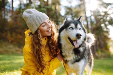 Şapkalı ve sarı paltolu güzel genç bir kadın parktaki çimlerde köpeğiyle yürüyor. Köpeğiyle dışarıda poz veren mutlu bir kadın. Arkadaşlık kavramı, tatil, yürüyüş.