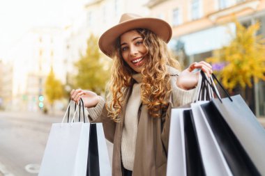 Alışveriş için çantalar, hediyeler, şehrin güneşli sokaklarında yürüyen ceketli ve şapkalı gülümseyen bir kadın. Ticaret kadını..