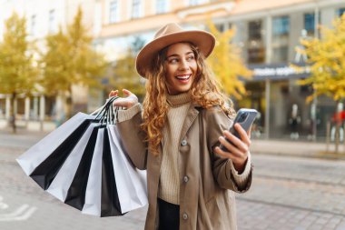 Alışveriş için çantalar, hediyeler, şehrin güneşli sokaklarında yürüyen ceketli ve şapkalı gülümseyen bir kadın. Ticaret kadını..