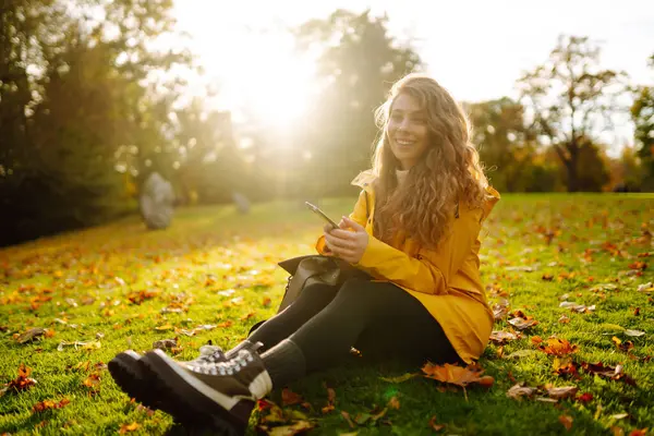 彼女の手のスマートフォンを持つ陽気な女性は 秋の公園で黄色い落ち葉の間のクリアに座っています 黄色いコートの女性は 秋の週末に時間を費やしています — ストック写真