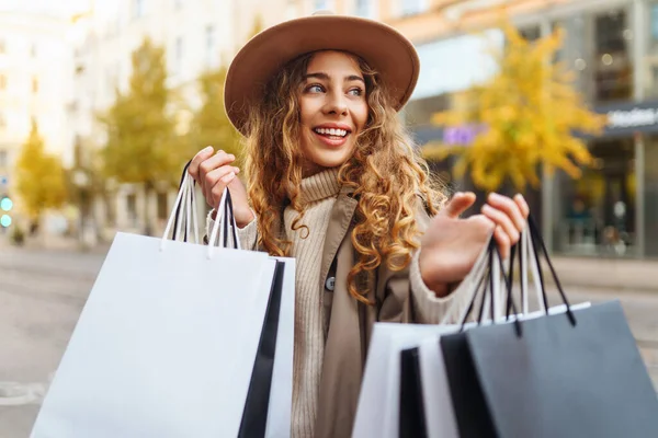 Alışveriş için çantalar, hediyeler, şehrin güneşli sokaklarında yürüyen ceketli ve şapkalı gülümseyen bir kadın. Ticaret kadını..