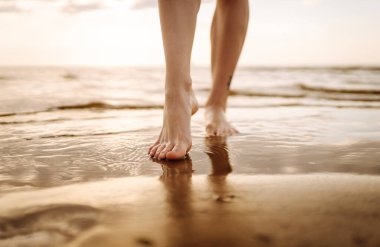 Sahilde deniz suyu ve kum dalgası üzerinde yürüyen genç bir kadının bacakları. Tatil, seyahat, özgürlük kavramı..