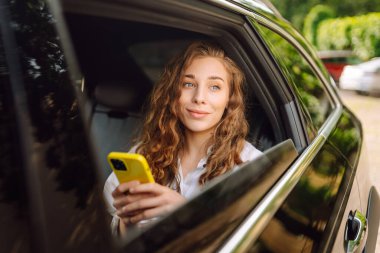 Gündelik giysiler içinde modern bir kadın arabanın arka koltuğunda oturur, kafasını çevirir, cep telefonu kullanır. İş, teknoloji ve seyahat kavramı.