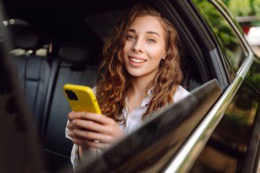 Gündelik giysiler içinde modern bir kadın arabanın arka koltuğunda oturur, kafasını çevirir, cep telefonu kullanır. İş, teknoloji ve seyahat kavramı.