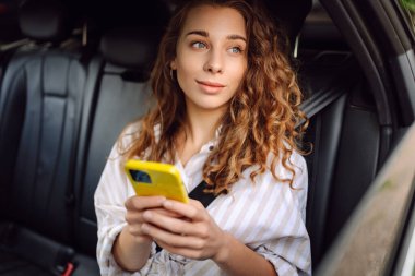 Gündelik giysiler içinde modern bir kadın arabanın arka koltuğunda oturur, kafasını çevirir, cep telefonu kullanır. İş, teknoloji ve seyahat kavramı.
