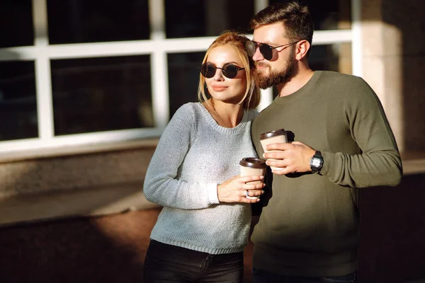 Stylish man and woman walk through the streets of the city and drink coffee together. Couple in love communicate while walking through the morning city. Concept of love, relationships, relaxation.