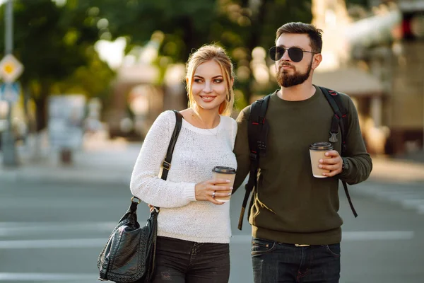 Stylish man and woman walk through the streets of the city and drink coffee together. Couple in love communicate while walking through the morning city. Concept of love, relationships, relaxation.