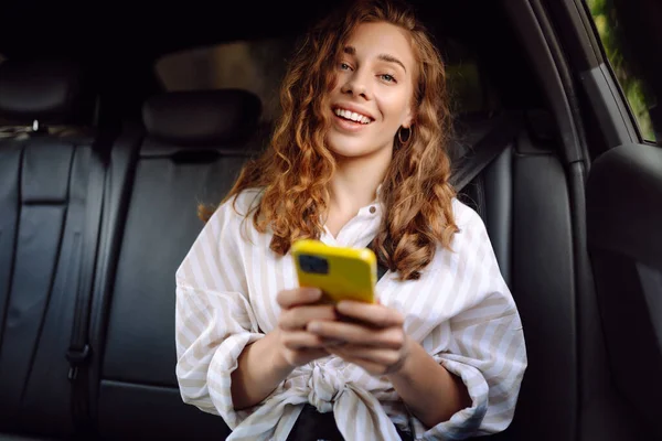 Gündelik giysiler içinde modern bir kadın arabanın arka koltuğunda oturur, kafasını çevirir, cep telefonu kullanır. İş, teknoloji ve seyahat kavramı.