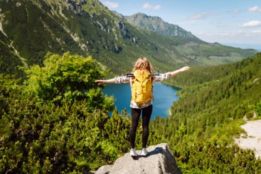 Parlak sarı sırt çantalı dişi turist dağ manzaralarının ortasında bir dağ gölünün yanında duruyor. Turizm, seyahat, yürüyüş kavramı.