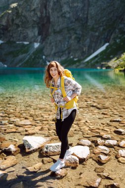 Parlak sarı sırt çantalı dişi turist dağ manzaralarının ortasında bir dağ gölünün yanında duruyor. Turizm, seyahat, yürüyüş kavramı.