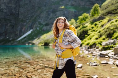 Parlak sarı sırt çantalı dişi turist dağ manzaralarının ortasında bir dağ gölünün yanında duruyor. Turizm, seyahat, yürüyüş kavramı.