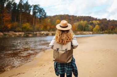 Sırt çantalı ve şapkalı gezgin bir kadın nehir kıyısında, doğada. Şık bir kadın güneşli bir günde hızlı nehrin yanında doğanın tadını çıkarır. Seyahat, doğa konsepti..