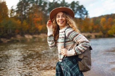 Sırt çantalı ve şapkalı gezgin bir kadın nehir kıyısında, doğada. Şık bir kadın güneşli bir günde hızlı nehrin yanında doğanın tadını çıkarır. Seyahat, doğa konsepti..