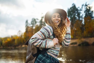 Sırt çantalı ve şapkalı gezgin bir kadın nehir kıyısında, doğada. Şık bir kadın güneşli bir günde hızlı nehrin yanında doğanın tadını çıkarır. Seyahat, doğa konsepti..