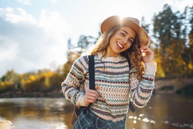 Sırt çantalı ve şapkalı gezgin bir kadın nehir kıyısında, doğada. Şık bir kadın güneşli bir günde hızlı nehrin yanında doğanın tadını çıkarır. Seyahat, doğa konsepti..