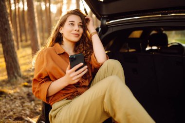 Ormanda bir arabanın bagajında şık giyinmiş genç kıvırcık bir kadın ve telefonu kullanıyor. Seyahat konsepti, aktif yaşam tarzı, hafta sonları.