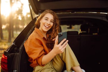Ormanda bir arabanın bagajında şık giyinmiş genç kıvırcık bir kadın ve telefonu kullanıyor. Seyahat konsepti, aktif yaşam tarzı, hafta sonları.