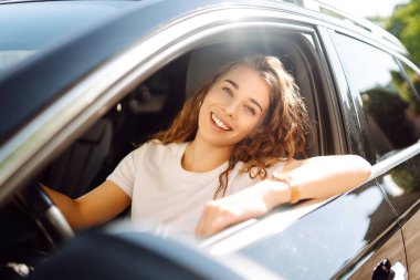 Gülümseyen kadın araba kullanıyor. Çekici bir kadın bir arabanın sürücü koltuğunda oturuyor. Açık hava portresi. Ulaşım konsepti, yaşam tarzı.