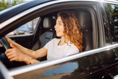 Gülümseyen kadın araba kullanıyor. Çekici bir kadın bir arabanın sürücü koltuğunda oturuyor. Açık hava portresi. Ulaşım konsepti, yaşam tarzı.