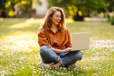 Parktaki çimlerin üzerinde oturmuş laptopla ders çalışan genç bir kadının portresi. Mutlu kadın çayırdaki güneşli havanın tadını çıkarıyor. Serbest çalışma kavramı, çevrimiçi öğrenme.