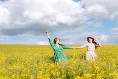 Şık giyinmiş kızlar yakut tohumu tarlasında birlikte yürüyorlar. İki kadın havanın tadını çıkarır, poz verir, çiçek açan kolza tohumlarının arasında eğlenir..