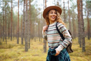 Sırt çantası ve şapkası olan aktif genç bir kadın ormanda güzel bir sonbahar gününün tadını çıkarıyor, ormanın çalılığını keşfediyor. Gezgin ormanda bir patika boyunca yürüyor..