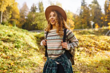 Sırt çantası ve şapkası olan aktif genç bir kadın ormanda güzel bir sonbahar gününün tadını çıkarıyor, ormanın çalılığını keşfediyor. Gezgin ormanda bir patika boyunca yürüyor..