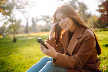 Sonbahar parkındaki açıklıkta elinde telefonla oturup sonbahar havasının tadını çıkaran tatlı bir kadın. İnsanların konsepti, rahatla. Yaşam biçimi.