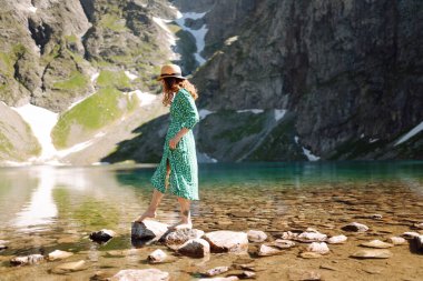 Elbiseli ve şapkalı genç bir kadın yüksek bir dağ gölünün manzarasının tadını çıkarıyor. Dağın arka planında poz veren romantik turist.