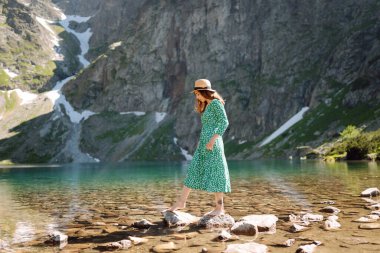 Elbiseli ve şapkalı genç bir kadın yüksek bir dağ gölünün manzarasının tadını çıkarıyor. Dağın arka planında poz veren romantik turist.