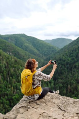 Young traveler with a yellow backpack is resting and taking pictures on the phone on a mountain cliff, admiring the landscape of the high Tatras, mountain range. Beautiful woman on top of the mountain clipart