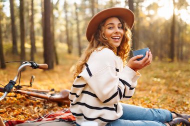 Şapkalı güzel kıvırcık kadın sonbahar ormanında termosla kırmızı bir mindere oturur. Kadın mini piknikte doğanın tadını çıkarıyor. Rahatlama kavramı, doğa..